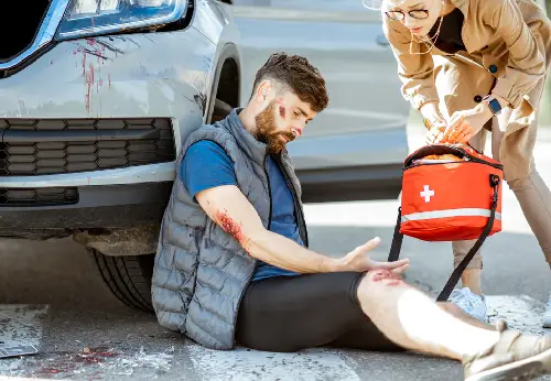 Injured man sitting beside a wrecked car as a woman approaches with a first aid kit to help
